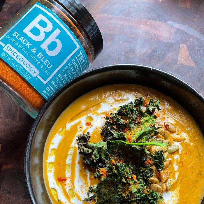Black & Bleu Roasted Squash Soup featuring roasted autumn squash, onions and garlic with a healthy shake of Black & Bleu garnished with crispy kale and pumpkin seeds in a bowl next to a jar of Spiceology Black & Bleu on a wooden background