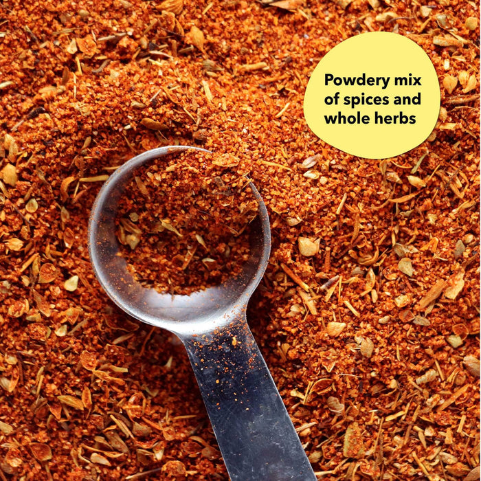 Overhead close-up of a dark reddish-brown spice blend with a silver measuring spoon; a callout label reads Powdery mix of spices and whole herbs