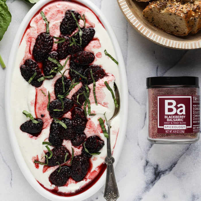 Blackberry Balsamic Roasted Blackberries with Whipped Feta featuring blackberries with a dusting of our Spiceology Blackberry Balsamic blend, a drizzle of honey and garnished with fresh basil in a white dish next to a jar of Spiceology Blackberry Balsamic on a marbled background