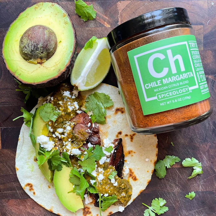 Chile Margarita Charred Salsa Verde featuring green tomatillos, yellow onion, jalapeno, garlic, cilantro and olive oil on a tortilla with avocado slices next to a jar of Spiceology Chile Margarita, avocado half and lime wedge on a wooden background