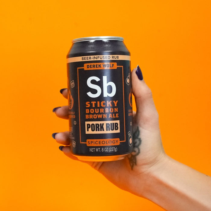 Hand holding a can of Spiceology Derek Wolf Sticky Bourbon Brown Ale Pork Rub against an orange background