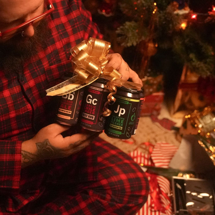 Person holding Derek Wolf Ultimate 6-Pack Beer-Infused Rub Sampler with a gold bow in front of a Christmas tree.