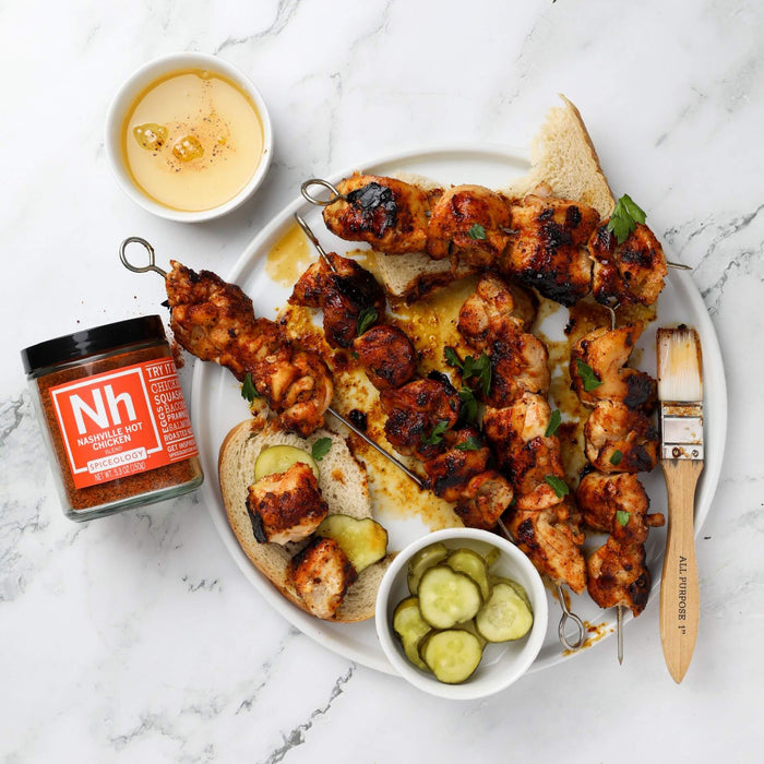 Overhead plate of grilled chicken skewers with pickles and a Spiceology Nashville Hot Chicken jar on a marble surface