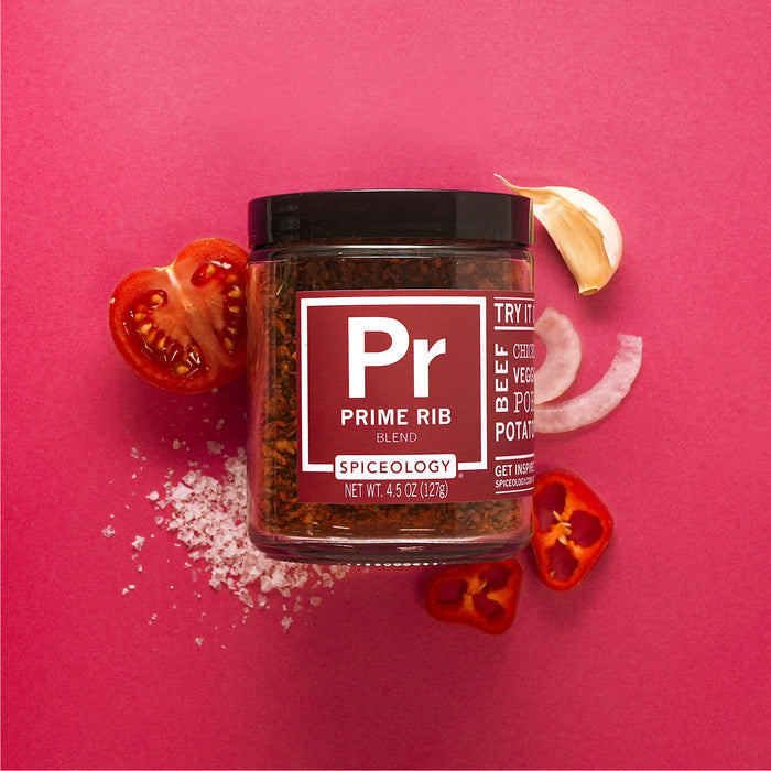 Jar of Spiceology prime rib rub on a bright red background with a clove of garlic, onion, tomato and red pepper slices