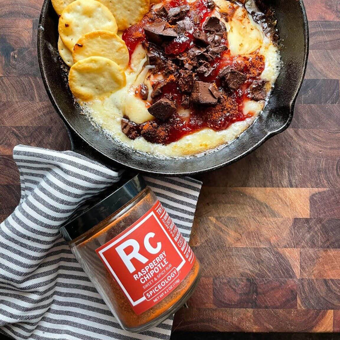 Raspberry Chipotle Baked Brie featuring wheel a brie, some jam, chocolate and crackers in a pan on a wooden surface next to a jar of Spiceology Raspberry Chipotle blend
