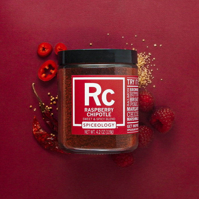 Lar of Spiceology raspberry chipotle sweet & spicy blend on a deep red background with red chili peppers, red raspberries, red peppers slices and sugar granules