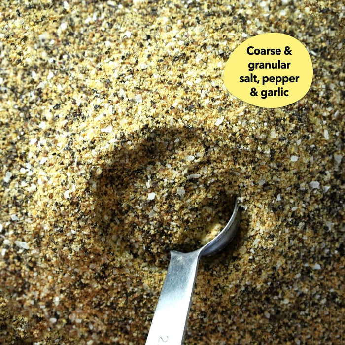 Overhead close-up of a coarse golden-green salt, pepper, and garlic blend with a silver measuring spoon; a callout reads Coarse and granular salt, pepper and garlic