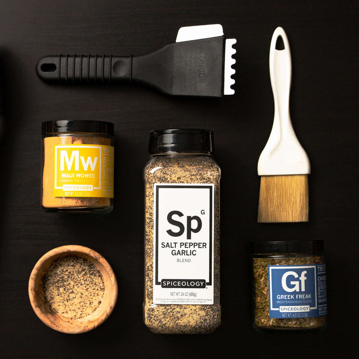 Flat lay of Spiceology Maui Wowee, Salt Pepper Garlic, and Greek Freak jars on an orange surface with a pastry brush and spreader