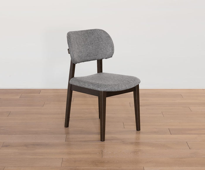 The Severin Dining Chair, featuring a dark gray finish, curved backrest, and plush cushioning, sits atop a light wood floor against a plain white wall—its dark wooden legs enhancing the mid-century charm.