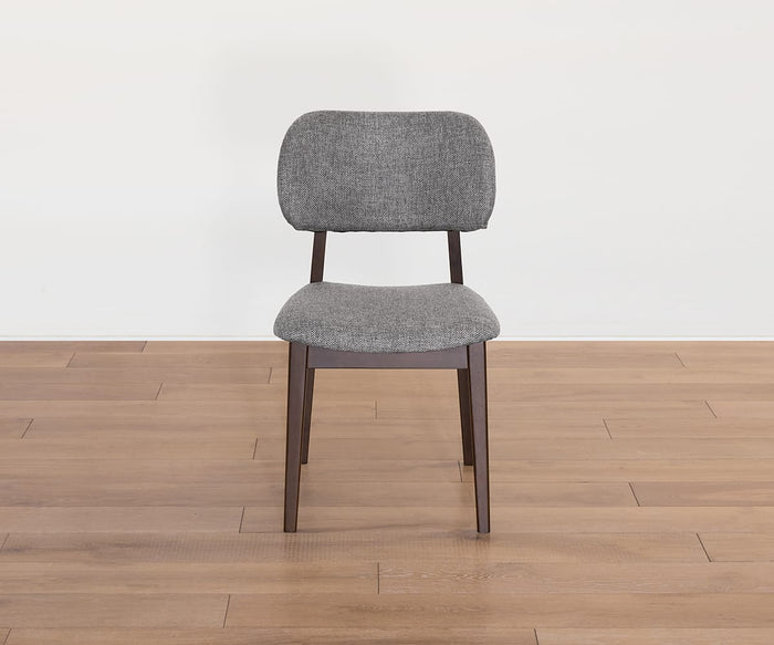 The Severin Dining Chair, featuring gray upholstery, a curved backrest, and dark rubberwood legs, is positioned on a light brown wooden floor against a plain white wall.