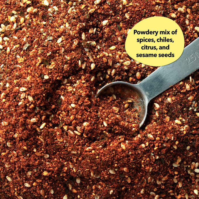 Overhead close-up of a warm reddish-orange spice blend with visible flakes and seeds and a silver measuring spoon; callout reads Powdery mix of spices, chiles, citrus, and sesame seeds