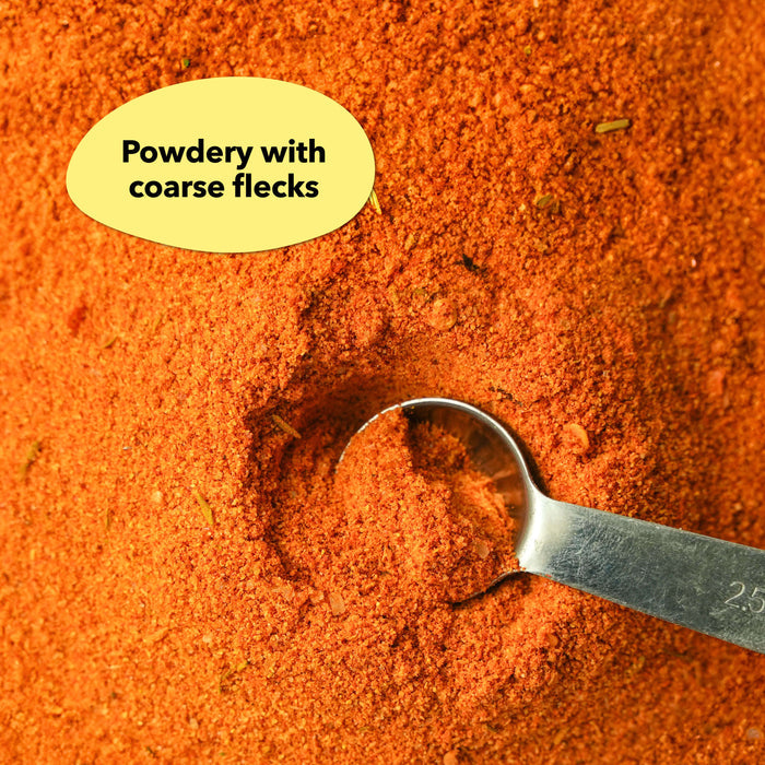 Overhead close-up of a reddish-orange spice blend with a silver measuring spoon resting in it; a callout label reads Powdery with coarse flecks