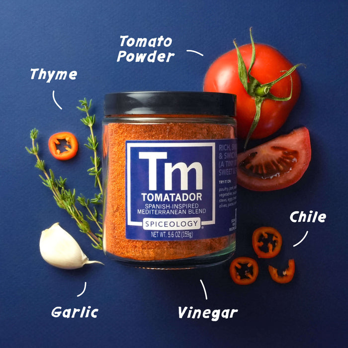 Spiceology Tomatador jar on a dark blue background with labeled ingredient callouts for Tomato Powder, Chile, Garlic, Vinegar, and Thyme; fresh tomatoes, garlic, and herbs surrounding the jar