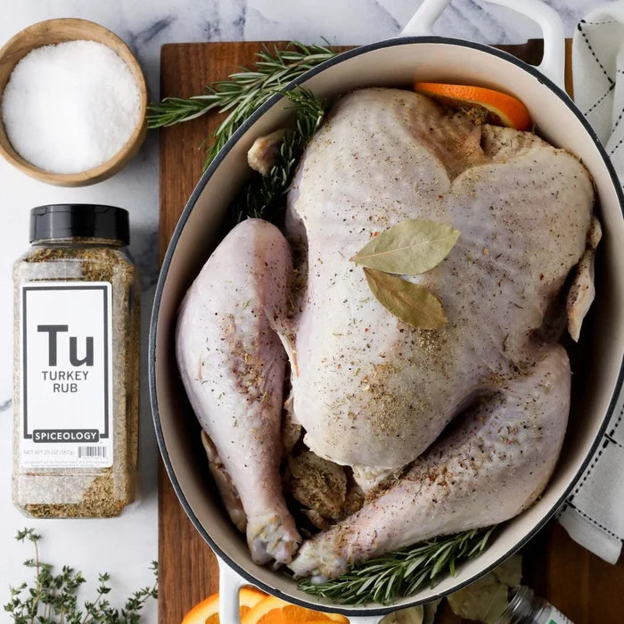 Overhead of a raw whole turkey in a roasting pan with fresh herbs and a Spiceology Turkey Rub jar on the side
